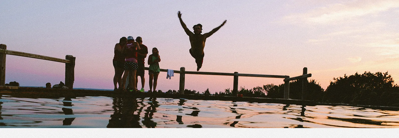 A teenager leaping off a dock into a lake at sunset while friends watch